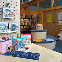 Zwei Tonieboxen 2 mit Tonieplay-Zubehör stehen auf einem Tisch in der Kinderbibliothek, umgeben von Toniefiguren, Büchern und gemütlichen Sitzmöbeln.